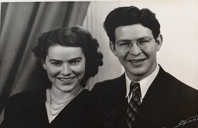 A black and white portrait of a young couple, a woman with curly hair and a necklace, and a man wearing glasses and a suit, both smiling.